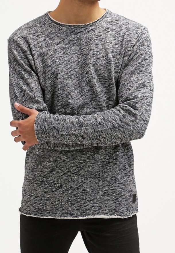Homme portant un pull en tricot gris chiné à manches longues avec un pantalon noir, les bras croisés sur la poitrine.