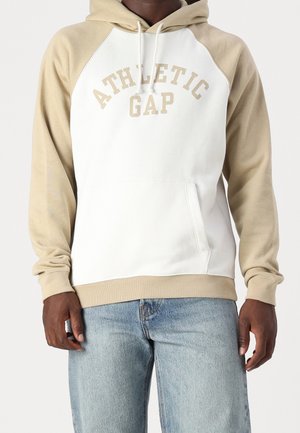 Mężczyzna w beżowo-białej bluzie z kapturem z napisem "ATHLETIC GAP" oraz w jasnoniebieskich dżinsach, stojący na tle jednolitego tła.