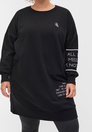 Osoba ubrana w czarną, oversize koszulę z długim rękawem, z białym tekstem na rękawie i przy dolnym brzegu, w połączeniu z ciemnoszarymi legginsami, na neutralnym tle.