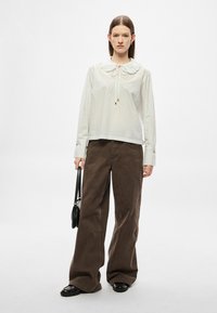 Biała bluzka w paski z dużym kołnierzem i ozdobą w postaci wiązania, w połączeniu z luźnymi brązowymi spodniami. Modelka trzyma małą czarną torebkę.