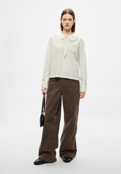 Blouse à rayures blanches avec un grand col et un détail de nœud, associée à un pantalon marron ample. Le mannequin tient un petit sac à main noir.