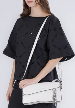 Femme portant un chemisier ajouré noir tenant un sac bandoulière en cuir blanc avec une fermeture éclair argentée et des breloques lettres décoratives.