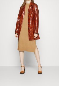 Brun läderjacka med glänsande textur, enfärgad design och fyra knappar, bärs över en ribbad beige klänning och plattformsklackar.