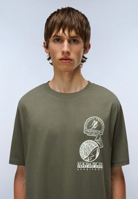 Camiseta de algodón verde oliva con tres logotipos impresos en blanco de diferentes tamaños, formas redondas y ovaladas, con cuello redondo y mangas cortas.