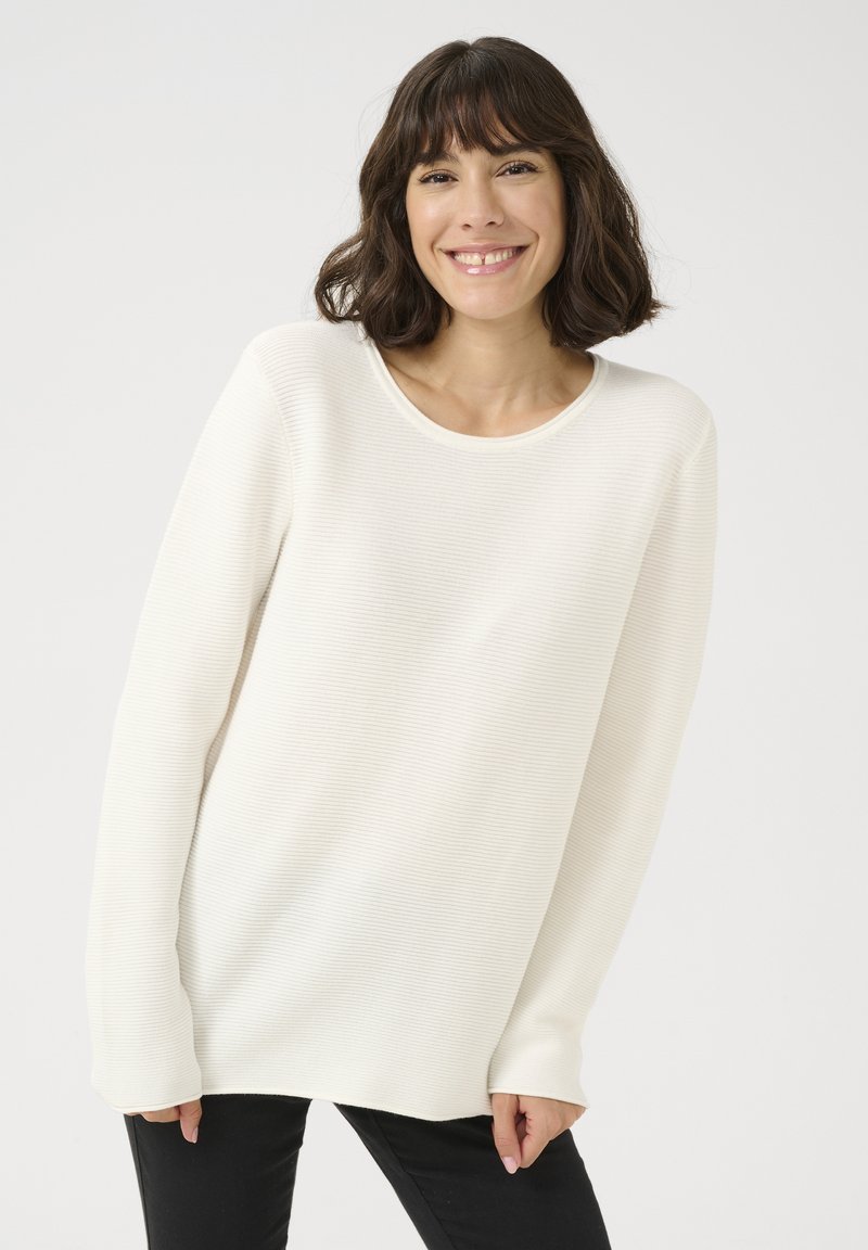 Maglione a maniche lunghe color crema con una texture a coste e scollatura rotonda, abbinato a pantaloni scuri e uno sfondo neutro.