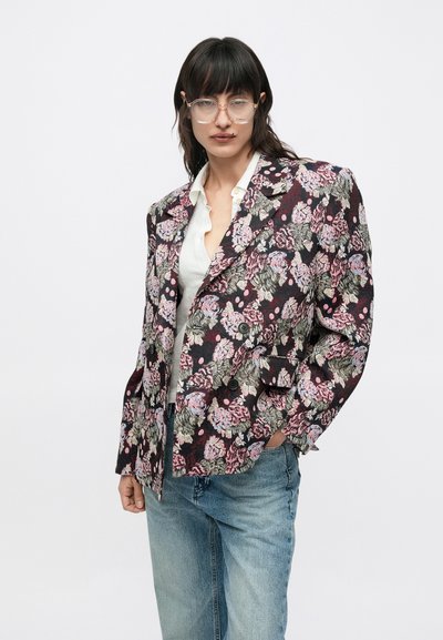 Δίπλο σακάκι με λουλουδάτο μοτίβο σε σκοτεινό φόντο, με ροζ και μπλε λουλουδένια σχέδια και μαύρα κουμπιά. Φοράται πάνω από λευκό πουκάμισο.
