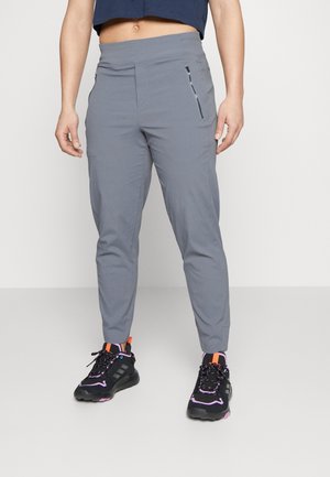 Personne portant un pantalon gris ajusté avec une poche latérale zippée et des chaussures de sport noires avec des accents violets et orange.
