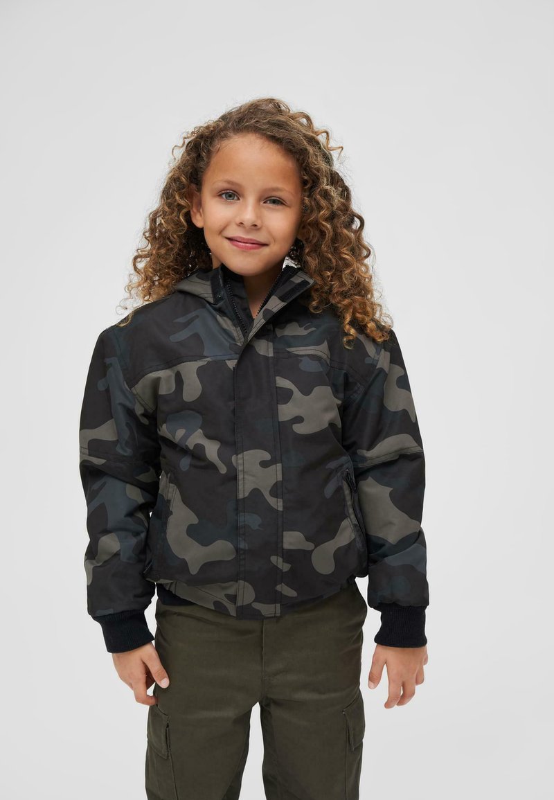Veste de camouflage noire et grise avec fermeture éclair à l'avant, capuche et poignets côtelés. Portée par-dessus un pantalon cargo olive, doté de plusieurs poches.