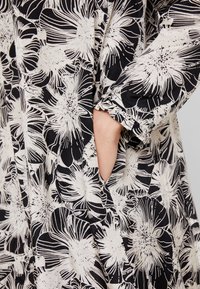 Czarny i biały materiał w kwiatowy wzór o luźnym kroju, z detalem kieszeni i gatheringiem na mankietach. Miękka tekstura.