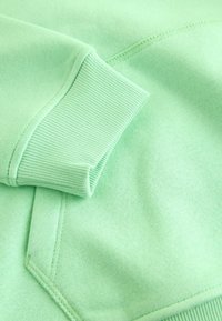 Light green sweatshirt sleeve with ribbed cuff resting near a stitched front pocket on the same fabric.