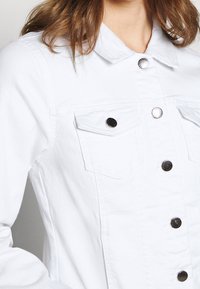 Chaqueta de mezclilla blanca con botones de metal plateado y bolsillos en el pecho, llevada por una persona con cabello castaño claro, cortada a mitad del torso.