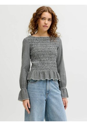 Tânără purtând o bluză din bumbac negru și alb cu talie strânsă, tiv și mâneci cu volănașe, asortată cu blugi de culoare bleu deschis, cu talie înaltă.