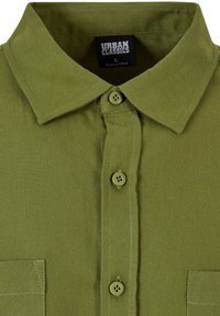 Camisa de color verde oliva con cuello, cierre de botones, dos bolsillos frontales y detalles de costura reforzada. La tela parece duradera y texturizada.