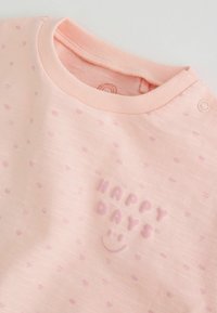 Világosrózsaszín babaing apróbb sötét rózsaszín pöttyökkel, patent gomb a vállon, és kiemelkedő "HAPPY DAYS" felirat egy mosolygós arc alatt.