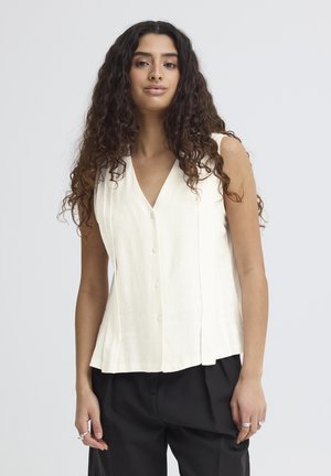 Mujer con cabello largo y rizado, que lleva una blusa blanca sin mangas con botones y pantalones negros, de pie frente a un fondo claro y liso.