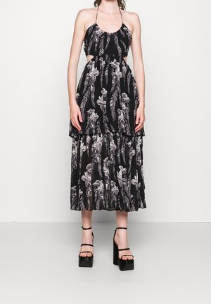 Femme portant une robe midi noire avec des imprimés floraux et plumes blancs, décolleté halter, découpes latérales et sandales à talons hauts noires.