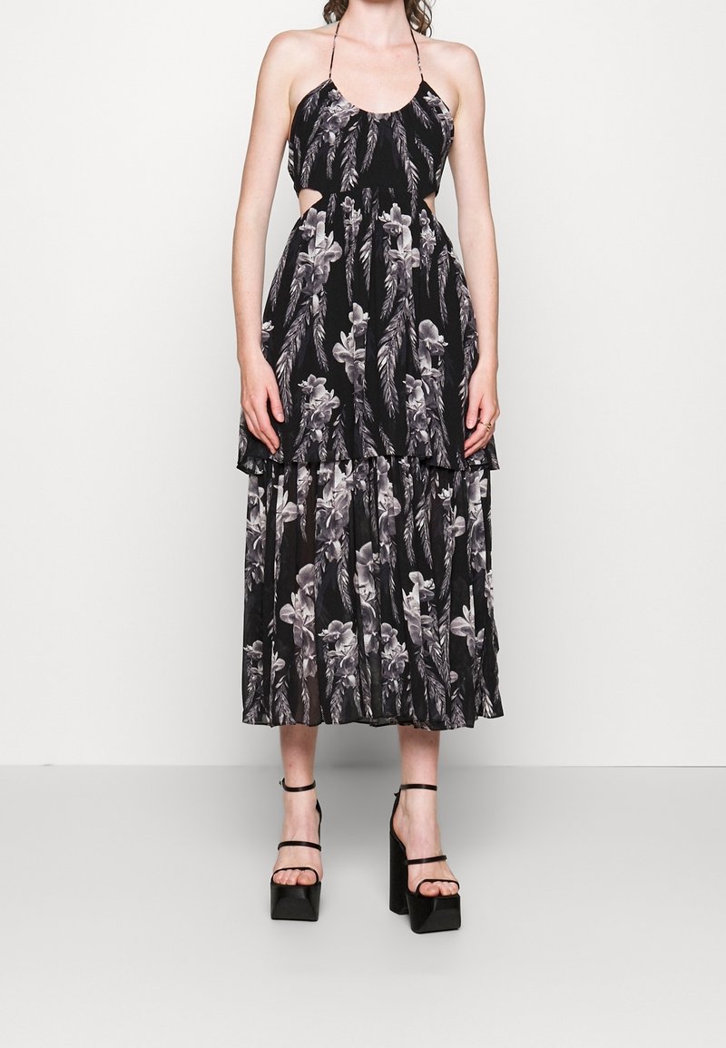 Nainen, jolla on päällä musta midi-mekko, jossa on valkoisia kukka- ja sulkapainatuksia, kauluksessa halkaisija, sivuleikkaukset ja mustat korkeat korkokengät.