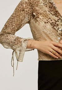 Blusa beige transparente con un estampado de leopardo marrón, presenta puños abotonados y lazos. Se lleva con un pantalón negro y joyas doradas en la mano.