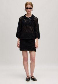 Schwarzes Strickoberteil mit Kragen und drei Knöpfen, kombiniert mit einem passenden Miniröckchen. Verfügt über ein Spitzenmuster und lockere Ärmel. Schwarze Schuhe.