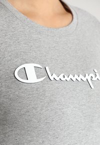 Camiseta de algodón gris con un destacado logo blanco de "Champion" en cursiva con un acento circular, cuello redondo acanalado y textura suave.