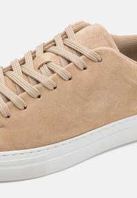 Beżowe zamszowe sneakersy z okrągłym noskiem, białą gumową podeszwą i płaskimi beżowymi sznurówkami. Charakteryzują się teksturowanym panelem pięty oraz minimalistycznym designem.