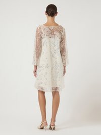 Γυναίκα με καστανά μαλλιά σε κότσο, φορώντας ένα διάφανο λευκό φόρεμα μέχρι το γόνατο με ανθισμένα και παγιέτα διακοσμητικά στοιχεία, και μπεζ παπούτσια με χαμηλό τακούνι, γυρισμένη προς τα πίσω.