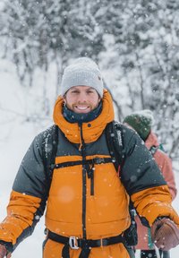 Bright orange and gray puffer jacket with a high collar, paired with a gray knitted beanie. Snow falls in a winter outdoor setting.