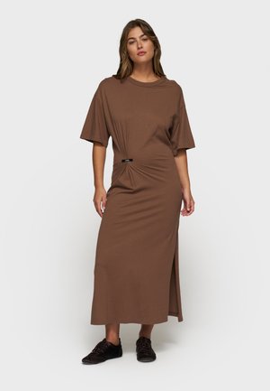 Donna che indossa un abito midi marrone con maniche corte, spacco laterale, dettaglio arricciato in vita e scarpe casual nere, in piedi su uno sfondo bianco.