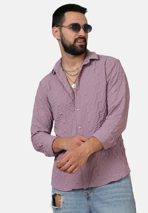 Camicia button-up di un leggero colore viola con un motivo testurizzato, maniche lunghe e colletto, abbinata a jeans strappati e collane sovrapposte.