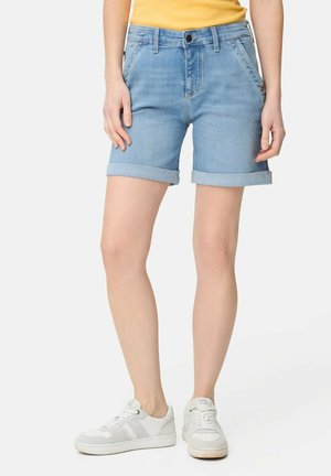 Osoba ubrana w jasnoniebieskie podwinięte szorty dżinsowe, białe trampki i żółtą koszulkę, stojąca na jednolitym tle.