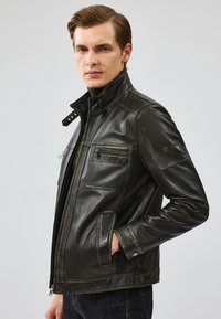 Veste en cuir noir avec un col haut, plusieurs poches et une fermeture éclair. Présente une texture lisse et des détails de conception angulaires.