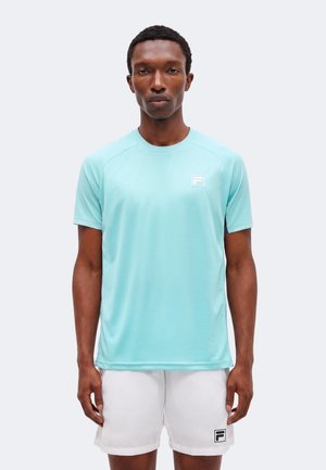 Homme portant un maillot de sport bleu clair à manches courtes et un short blanc avec logo, debout devant un fond clair uni.