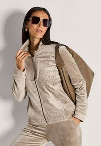 Vrouw in een beige velours trainingspak met subtiel luipaardpatroon, die een bruine leren tas over haar schouder houdt en grote zonnebrillen draagt.