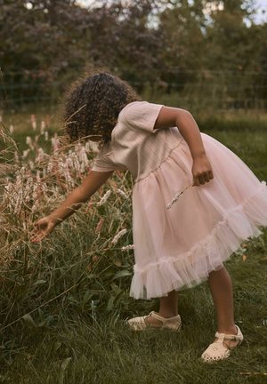 Mladé dievča v svetloružových šatách a pletených topánkach sa ohýba, aby nazbieralo divé kvety na trávnatej lúke s lesom v pozadí.