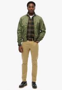 Chaqueta bomber verde sobre una camisa de cuadros en tonos amarillos, negros y azul marino. Chinos beige y zapatos negros con suelas de color marrón claro.