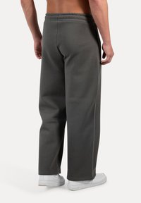 Mies seisoo paljain jaloin, päällään löysät tummanharmaat verkkarit ja valkoiset lenkkarit, shown selältä yksinkertaista valkoista taustaa vasten.