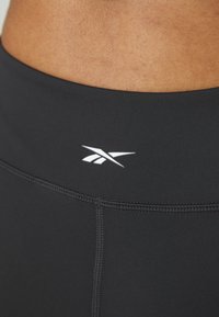 Leggings sportivi neri realizzati in tessuto elastico, con una texture liscia e un logo bianco in vita. Le cuciture sono visibili.