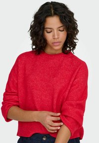 Pull rouge avec un tricot texturé, encolure ronde et manches retroussées. Le tissu semble doux, avec une coupe décontractée et légèrement raccourcie.