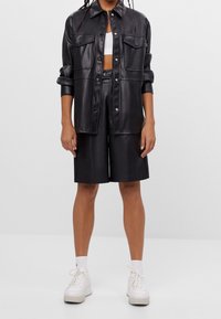 Person som bär en svart faux läder skjorta med knappar och matchande knälånga shorts, en vit crop top, vita strumpor och vita sneakers.