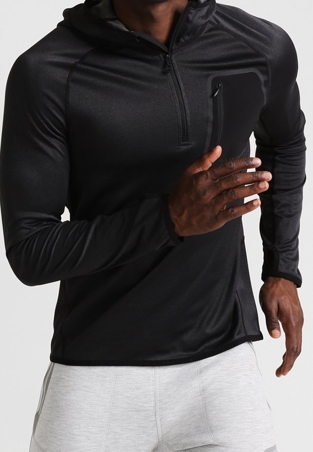 Mann, der in einem schwarzen Langarm-Zip-Hoodie mit Brusttasche und hellgrauen Sportshorts vor einem schlichten Hintergrund läuft.