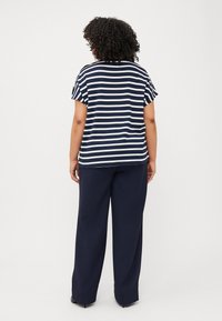 Modell med krøllete hår vender bort, iført en stripete topp i marineblått og hvitt med korte ermer og marineblå bukser, mot en ensfarget hvit bakgrunn.