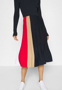 Jupe plissée avec des panneaux verticaux en noir, rouge, beige et blanc. Le tissu est côtelé avec une texture lisse. Associée à des baskets blanches.