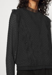 Musta paita, jossa on tekstuurikangas, ja joka on varustettu pilkkuilla sekä pitsiyksityiskohdilla olkapäillä ja kaula-aukossa. Pitkät hihat, joissa on frillireunat.