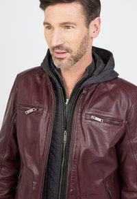 Burgunderfarbene Lederjacke mit zwei Reißverschlussbrusttaschen, silberner Hardware und einem grauen, Reißverschluss-Hoodie darunter, der eine strukturierte Oberfläche aufweist.