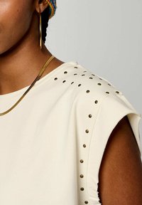 Haut crème à manches courtes avec des clous dorés le long de l'épaule, un col rond et un tissu doux et texturé. Accentué avec des bijoux.