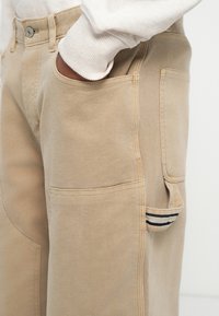 Pantaloni in cotone beige con taglio dritto, dotati di tasche laterali e una piccola tasca a toppa con un accento a righe vicino all'ancore.