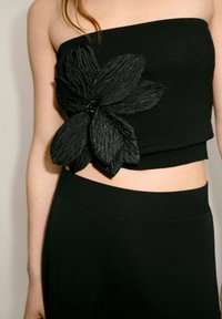 Top senza spalline lavorato a maglia nera con una decorazione in tessuto a forma di fiore nero con un centro di perline. Abbinato a un pantalone nero aderente.