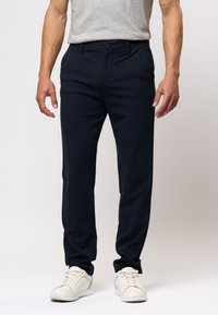 Férfi sötétkék slim-fit nadrágot, szürke pólót és fehér sportcipőt visel, egy sima világos háttér előtt.