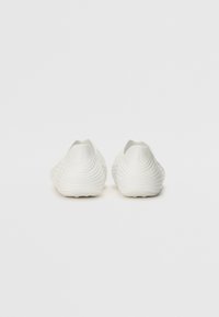 Scarpe bianche slip-on con una superficie testurizzata ondulata, tacco arrotondato e design minimalista, realizzate in un materiale liscio simile alla gomma.