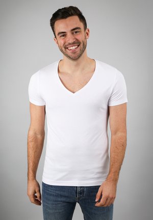 Smiling man with short dark hair wearing a white V-neck T-shirt and blue jeans, standing against a plain gray background.